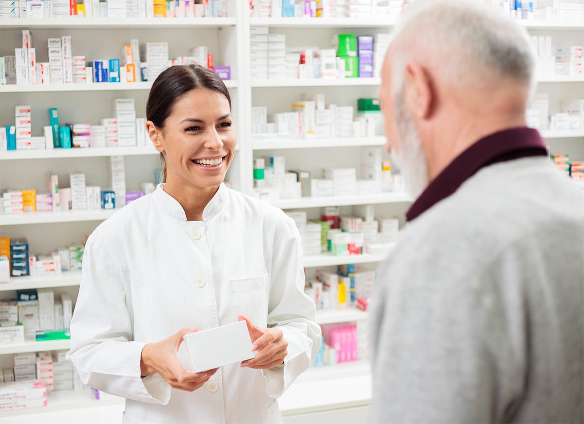 A smiling pharmacist helping a senior customer with a shelf full of medications behind her.