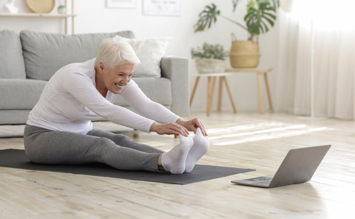 A senior woman stretching on a yoga mat, indoors, in front of her computer.