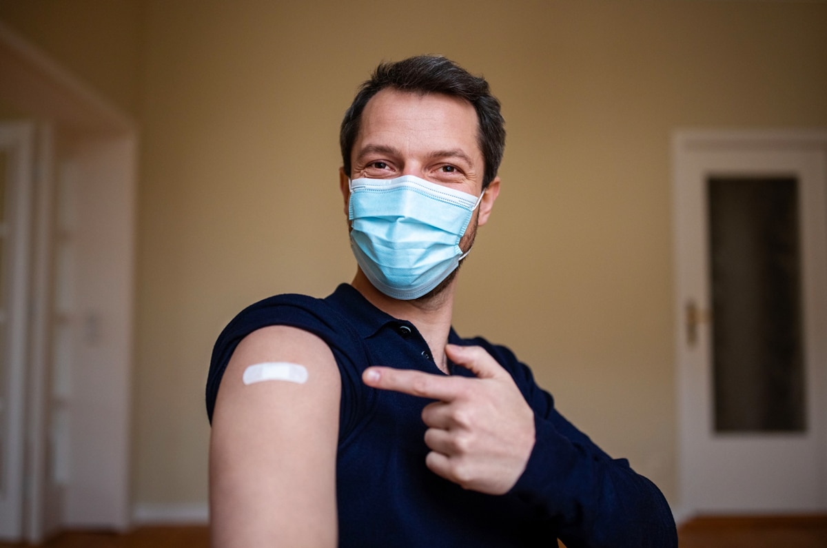 A man wearing a mask, pointing to a bandaid on his upper arm where he recently received a shot