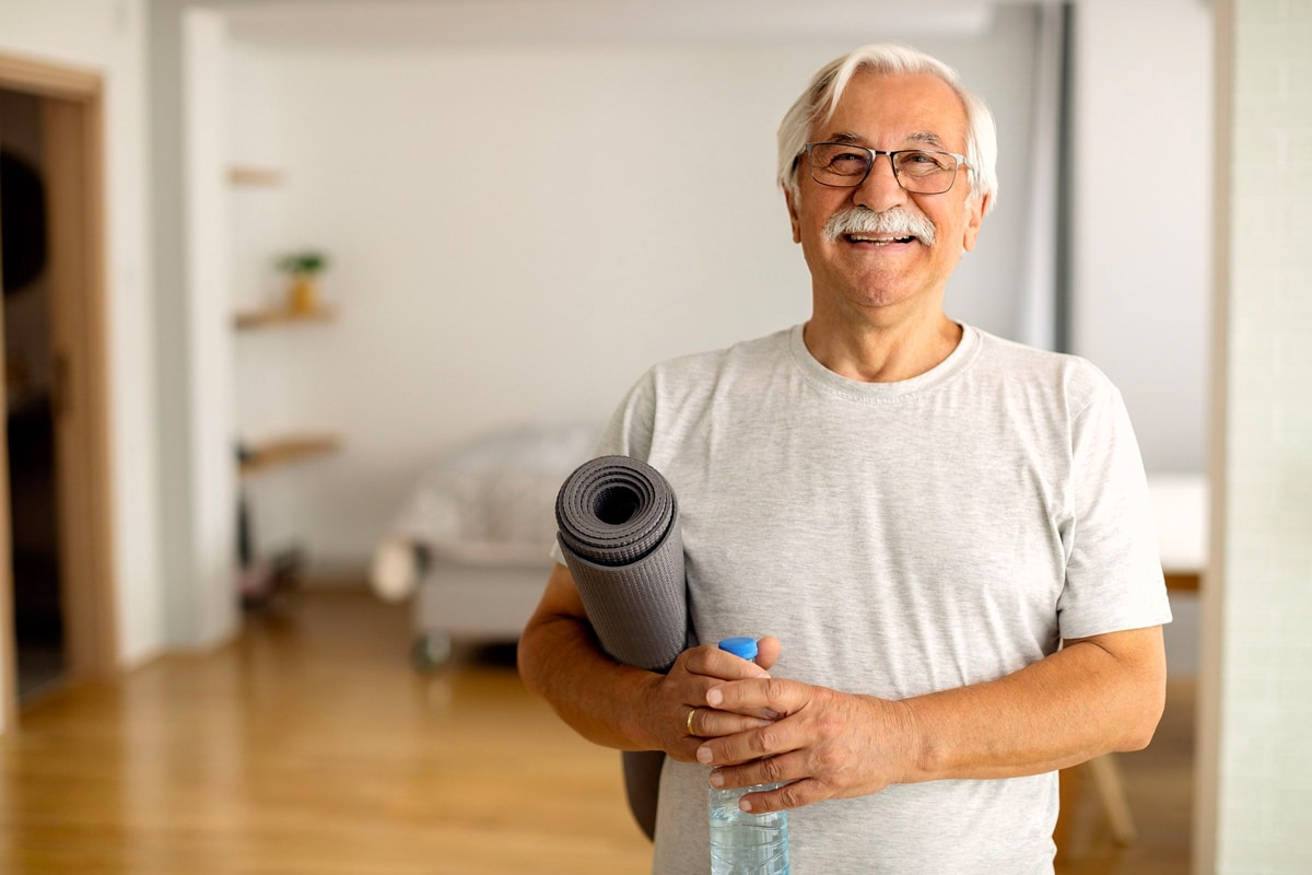 A smiling senior man holding a yoga mat under his arm, and a water bottle in his hands.