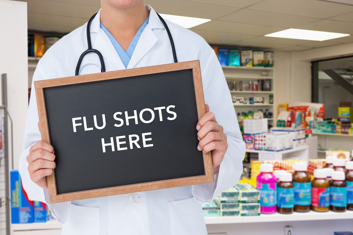 A pharmcist standing in front of medications, holding a small chalkboard sign that reads "Flu Shots Here."