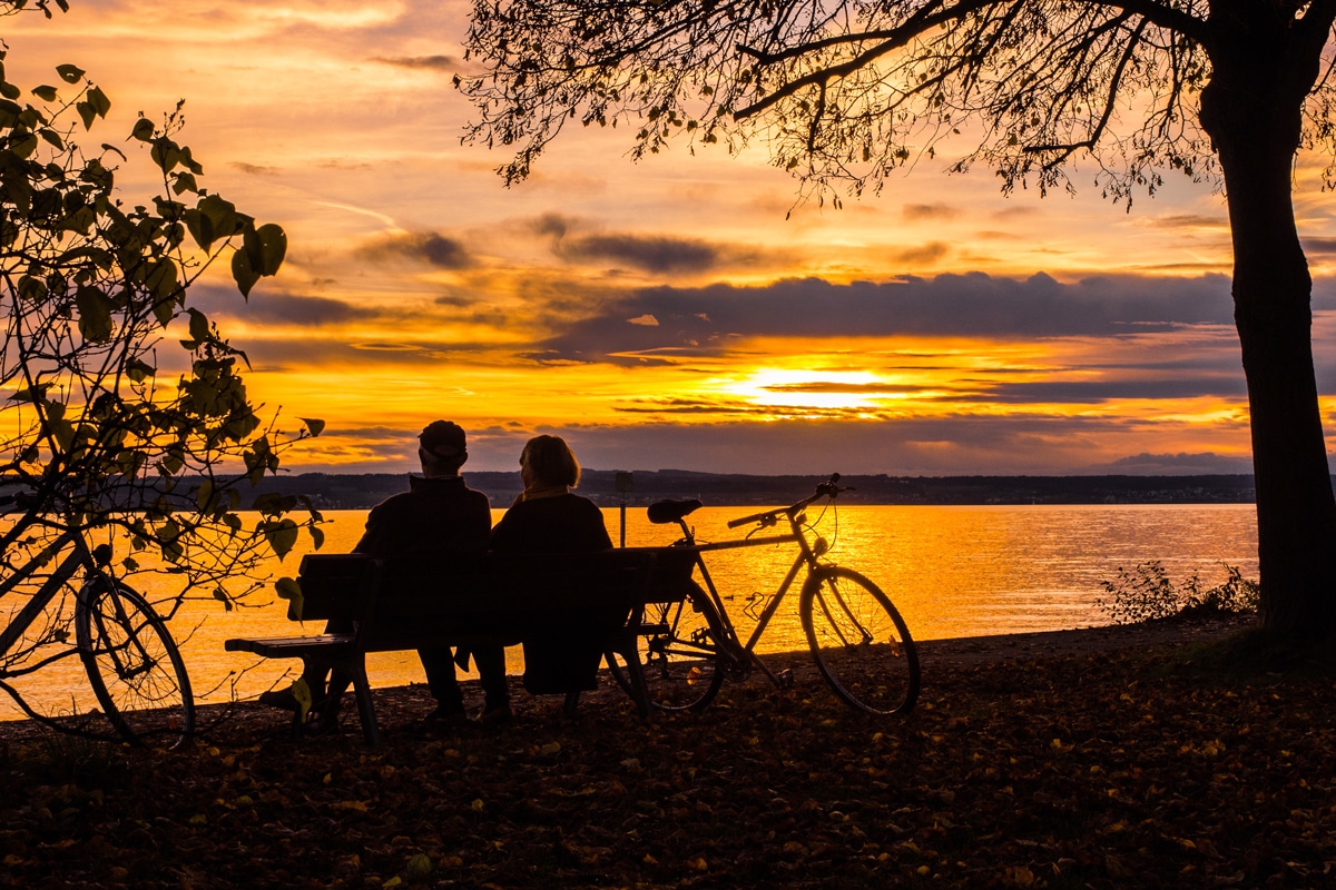 A senior couple sitting on a bench beside a lake at sunset.