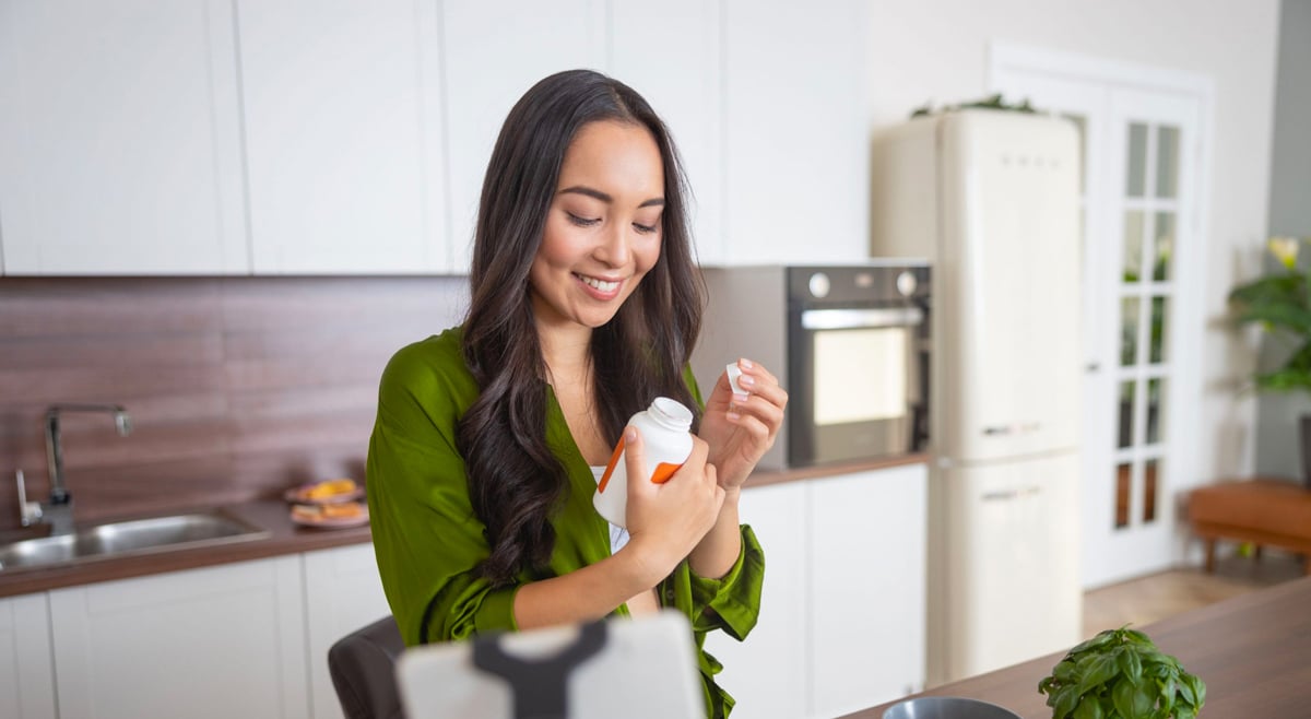 A smiling woman open a bottle of medication in her kitchen.
